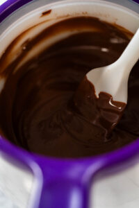 Rich Chocolate Royal Icing for Decorating - Arina Photography Melted Chocolate in Melting Pot with Spatula Description: Close-up view of melted chocolate being stirred with a spatula in a purple melting pot, achieving a smooth consistency for Chocolate Royal Icing.